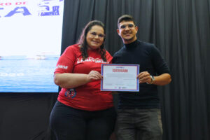 Fotografia de entrega de prêmio/certificado em evento acadêmico. Em plano médio, duas pessoas estão no palco em frente a uma cortina escura e uma tela de projeção à esquerda. À esquerda: Uma mulher de óculos e camiseta vermelha de evento acadêmico segura o certificado junto ao participante. À direita: Breno Brasileiro, mestrando em Neuroengenharia pelo ISD, vestindo blusa preta de gola alta e óculos, sorri para a câmera enquanto segura o certificado de premiação/reconhecimento.
