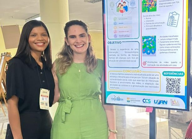 A imagem mostra duas mulheres sorridentes em pé lado a lado, posando ao lado de um pôster acadêmico fixado em um suporte preto, em um ambiente interno e iluminado. A mulher à esquerda tem pele morena, cabelos pretos lisos na altura dos ombros e veste uma camisa preta de mangas curtas e calça preta. Ela usa um crachá pendurado no pescoço. A mulher à direita tem pele clara, cabelos-castanho claros ondulados caindo sobre os ombros e veste um macacão verde-claro sem mangas, com uma faixa amarrada na cintura. Ela está posando com o braço em volta da mulher ao seu lado. Do lado direito da imagem, está o pôster científico impresso com fundo branco, detalhes em azul e verde e vários blocos de texto, gráficos e ilustrações.