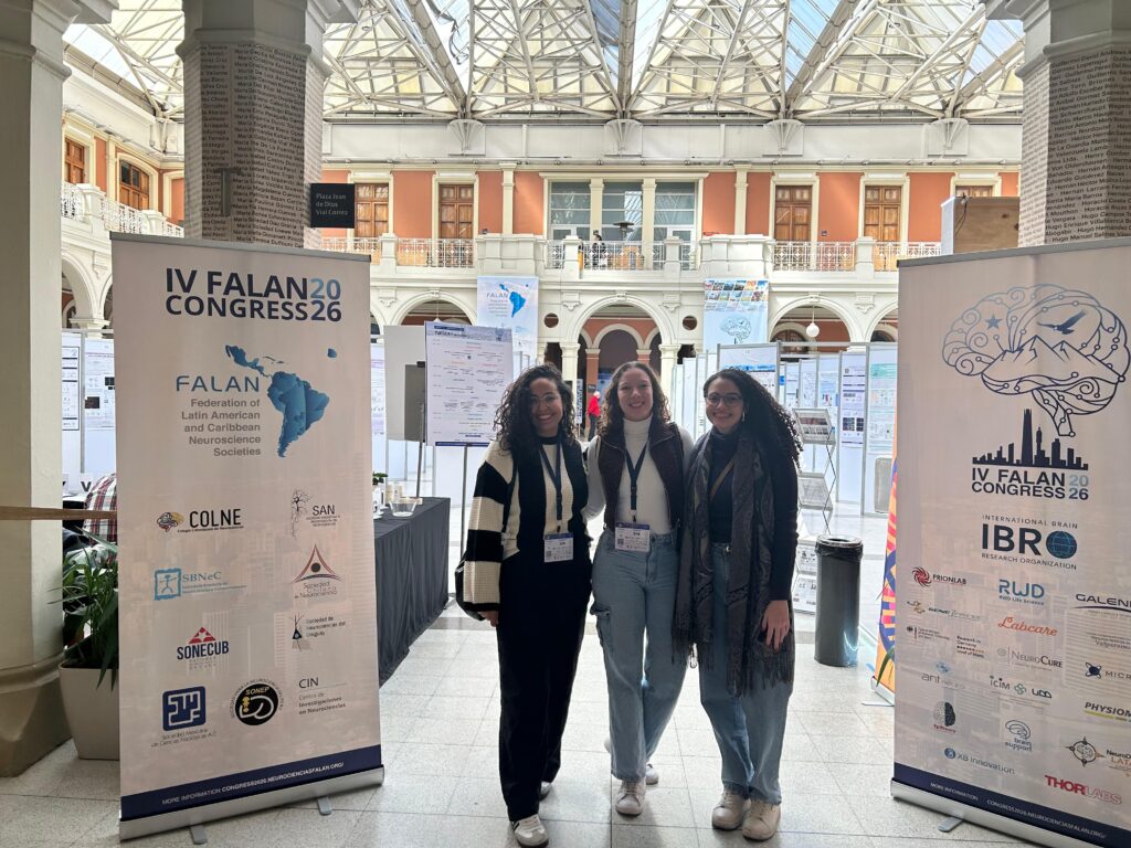 Fotografia em plano inteiro de três pesquisadoras (estudantes e egressas do ISD) posicionadas lado a lado durante o IV FALAN Congress. Elas sorriem para a câmera no saguão de um prédio histórico iluminado por luz natural, cercadas por banners informativos do evento internacional e painéis de trabalhos científicos. Foto: Acervo pessoal dos pesquisadores.