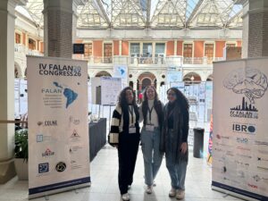 Fotografia em plano inteiro de três pesquisadoras (estudantes e egressas do ISD) posicionadas lado a lado durante o IV FALAN Congress. Elas sorriem para a câmera no saguão de um prédio histórico iluminado por luz natural, cercadas por banners informativos do evento internacional e painéis de trabalhos científicos. Foto: Acervo pessoal dos pesquisadores.