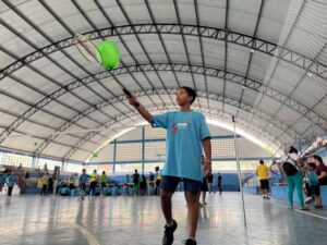 A imagem mostra um menino em primeiro plano, visto de um ângulo baixo, praticando uma atividade esportiva no centro de um ginásio coberto. O menino tem pele morena, cabelos curtos e escuros, e veste uma camiseta azul-clara e short jeans escuro. Ele segura uma raquete de badminton levantada com a mão direita e rebate um balão/bexiga verde-alface que substitui a peteca. O ginásio possui estrutura metálica no teto curvo, chão de quadra poliesportiva e paredes brancas com detalhes em azul. Ao fundo, em segundo plano, várias pessoas e crianças participam de outras atividades e vivências esportivas organizadas na quadra e nas arquibancadas laterais. Texto contido na estampa da camiseta do menino (verbatim): FESTIVAL PARALÍMPICO
