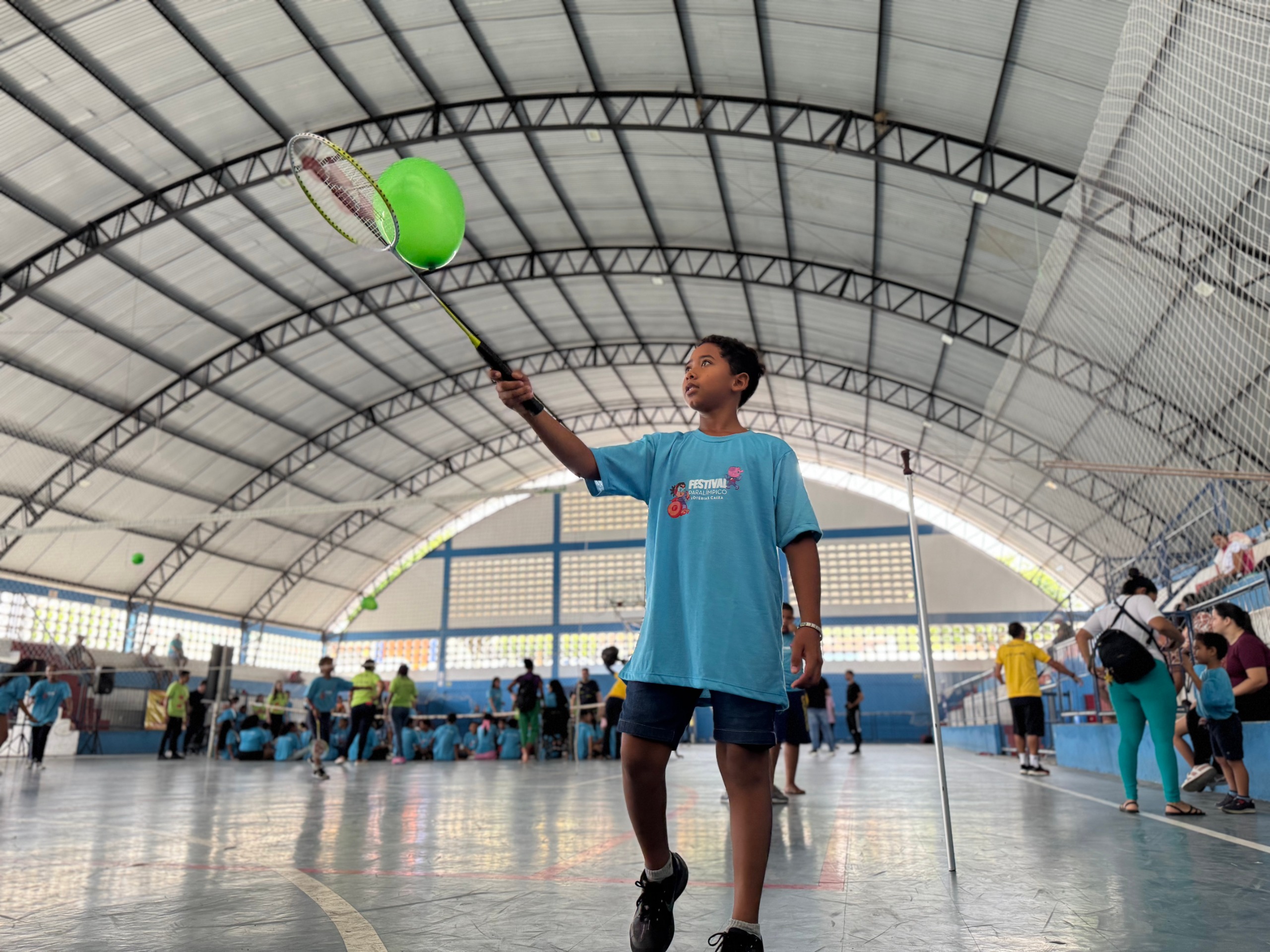 A imagem mostra um menino em primeiro plano, visto de um ângulo baixo, praticando uma atividade esportiva no centro de um ginásio coberto. O menino tem pele morena, cabelos curtos e escuros, e veste uma camiseta azul-clara e short jeans escuro. Ele segura uma raquete de badminton levantada com a mão direita e rebate um balão/bexiga verde-alface que substitui a peteca. O ginásio possui estrutura metálica no teto curvo, chão de quadra poliesportiva e paredes brancas com detalhes em azul. Ao fundo, em segundo plano, várias pessoas e crianças participam de outras atividades e vivências esportivas organizadas na quadra e nas arquibancadas laterais. Texto contido na estampa da camiseta do menino (verbatim): FESTIVAL PARALÍMPICO