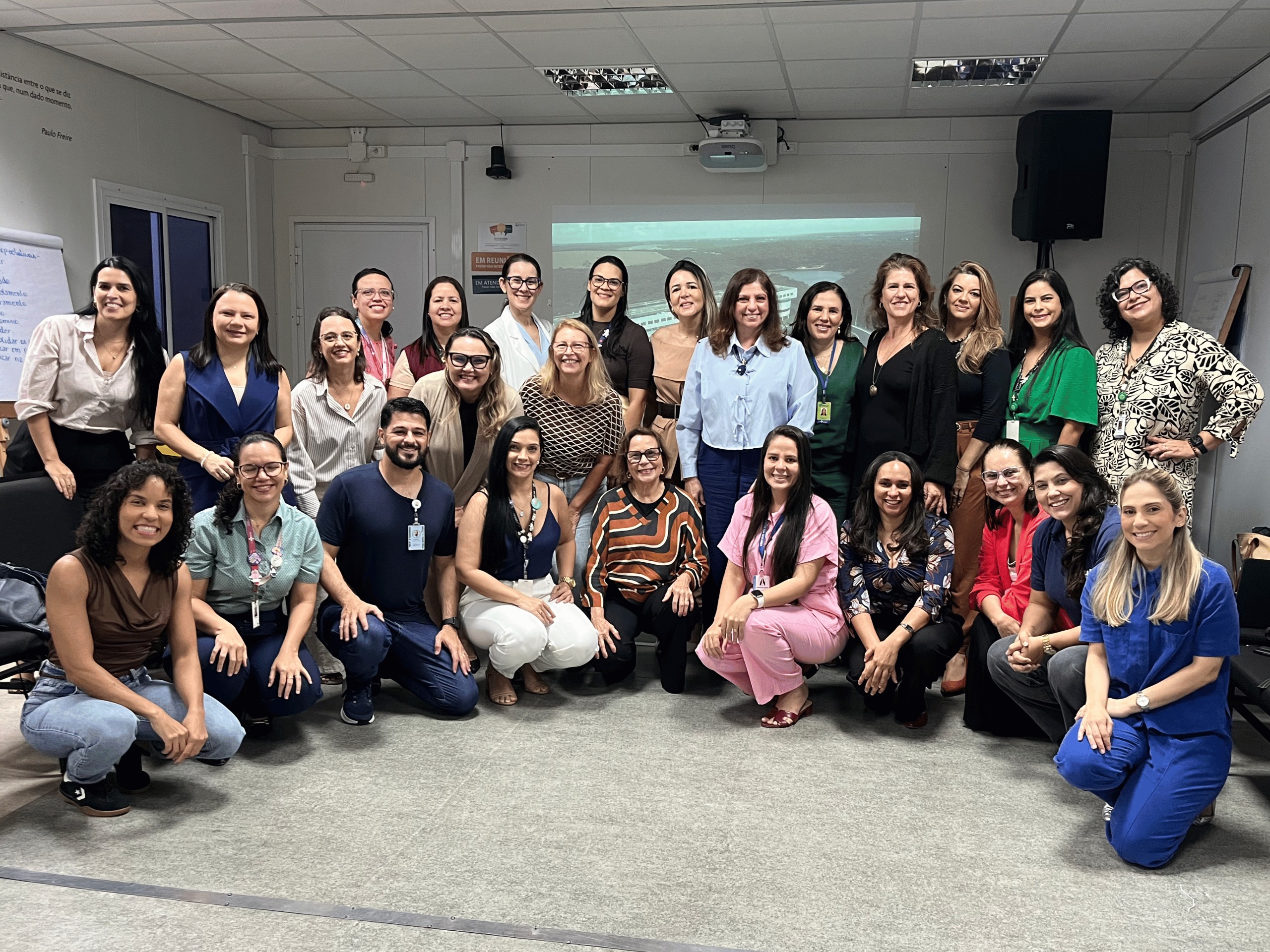 Fotografia em grupo reunindo vinte e quatro profissionais de saúde da atenção básica do Rio Grande do Norte ao final de uma oficina de capacitação. Os participantes aparecem sorridentes dispostos em duas fileiras, uma parte em pé ao fundo e outra ajoelhada ou agachada à frente. O grupo veste roupas casuais e trajes de trabalho em tons variados, posando no centro de uma sala de reuniões com projetor e tela ao fundo. Foto: Ascom ISD.