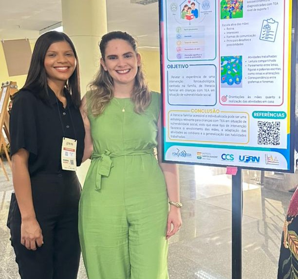 A imagem mostra duas mulheres sorridentes em pé lado a lado, posando ao lado de um pôster acadêmico fixado em um suporte preto, em um ambiente interno e iluminado. A mulher à esquerda tem pele morena, cabelos pretos lisos na altura dos ombros e veste uma camisa preta de mangas curtas e calça preta. Ela usa um crachá pendurado no pescoço. A mulher à direita tem pele clara, cabelos-castanho claros ondulados caindo sobre os ombros e veste um macacão verde-claro sem mangas, com uma faixa amarrada na cintura. Ela está posando com o braço em volta da mulher ao seu lado. Do lado direito da imagem, está o pôster científico impresso com fundo branco, detalhes em azul e verde e vários blocos de texto, gráficos e ilustrações.