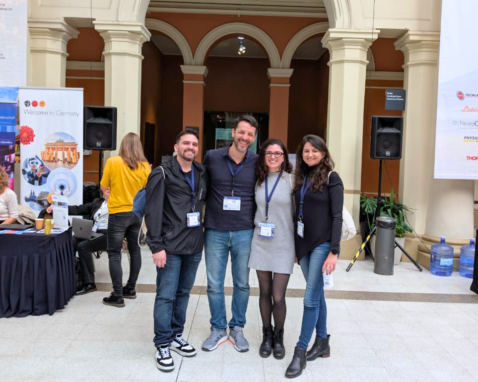 Fotografia em plano inteiro de quatro professores pesquisadores do ISD reunidos no Chile para um evento de neurociências. O grupo é composto por dois homens e duas mulheres que sorriem abraçados no pátio interno de um edifício de arquitetura clássica com arcos e colunas. Ao fundo, veem-se caixas de som, estandes e outros participantes do evento. Foto: Acervo pessoal dos pesquisadores.