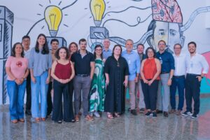 Fotografia em plano inteiro mostrando um grupo de quinze pessoas, homens e mulheres, lado a lado em uma sala iluminada. Os integrantes vestem trajes casuais e esporte fino, muitos deles usando crachás de identificação pendurados no pescoço. Ao fundo, a parede branca exibe um mural ilustrado em estilo grafite com lâmpadas acesas, circuitos elétricos, números binários e o desenho de um rosto com a cabeça aberta revelando o cérebro conectado a fios.