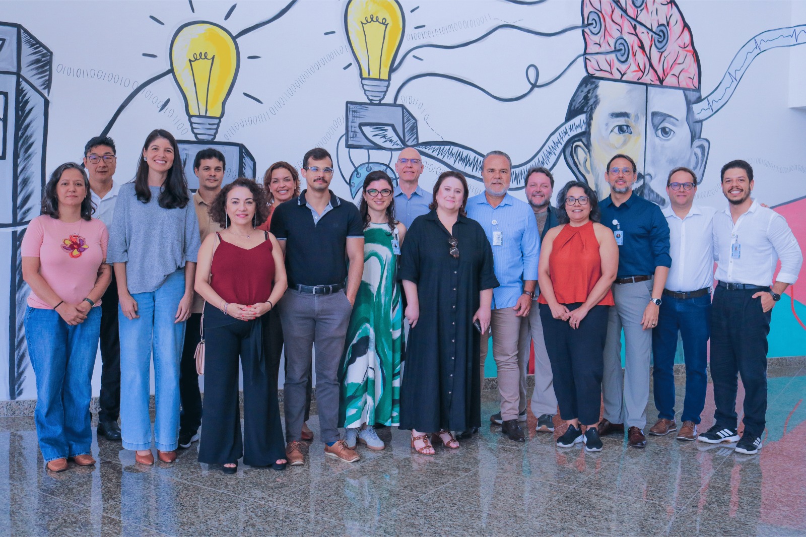 Fotografia em plano inteiro mostrando um grupo de quinze pessoas, homens e mulheres, lado a lado em uma sala iluminada. Os integrantes vestem trajes casuais e esporte fino, muitos deles usando crachás de identificação pendurados no pescoço. Ao fundo, a parede branca exibe um mural ilustrado em estilo grafite com lâmpadas acesas, circuitos elétricos, números binários e o desenho de um rosto com a cabeça aberta revelando o cérebro conectado a fios.