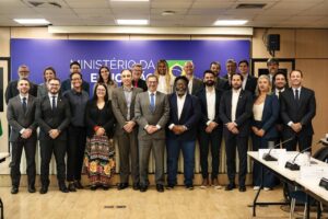 Fotografia em plano aberto de um grupo de cerca de vinte pessoas posando em pé em uma sala de reuniões ou auditório. Ao fundo, há um painel azul com a inscrição parcial "MINISTÉRIO DA EDUCAÇÃO" e a bandeira do Brasil. As pessoas, de diversos gêneros, idades e aparências, vestem roupas formais e casuais-formais (ternos, camisas e blazers) e sorriem levemente para a câmera. Em primeiro plano, mesas de reunião com microfones aparecem nas laterais inferior e direita.