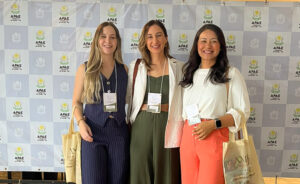 A imagem apresenta três mulheres posicionadas lado a lado e sorrindo em frente a um painel de fundo quadriculado com a marca da "APAE Campina Grande - PB". À esquerda, uma mulher de cabelos loiros e longos veste um conjunto risca de giz azul-marinho sem mangas. Ao centro, a mulher de blazer listrado claro e macacão verde segura uma bolsa tiracolo. À direita, uma mulher de cabelos pretos veste blusa branca e calça coral. As mulheres das extremidades carregam sacolas ecológicas de pano com a impressão "XXII SEMINÁRIO CONHECENDO PARA INTERVIR". Todas usam crachás de identificação do evento pendurados no pescoço.