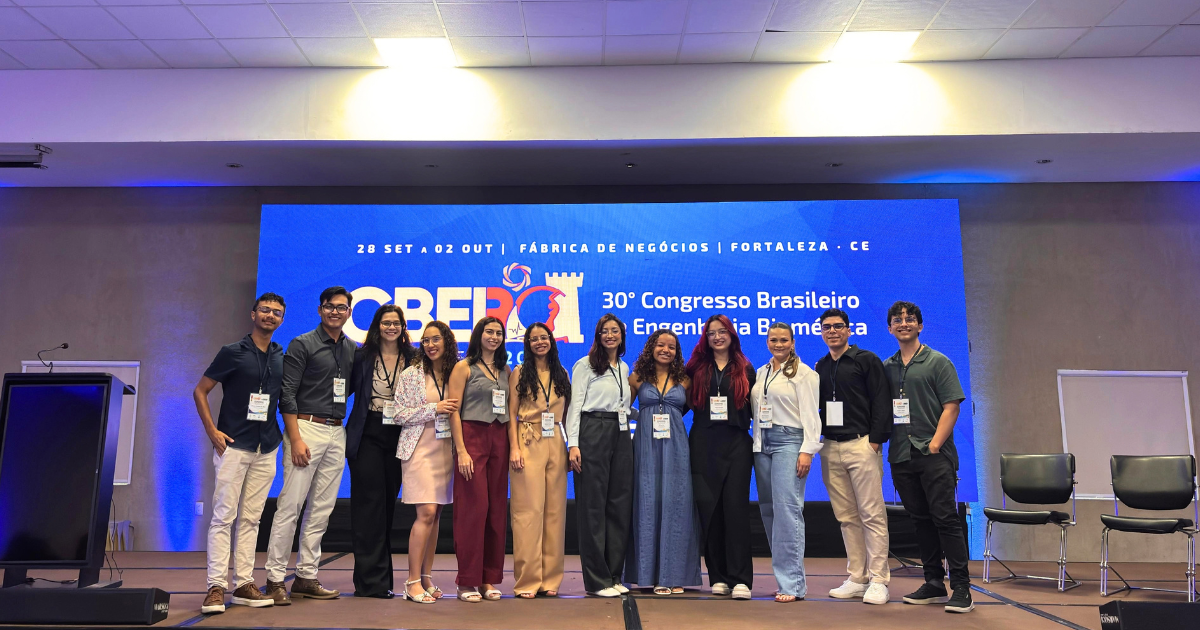 A imagem é uma fotografia em plano geral e formato horizontal, tirada no palco de um auditório ou salão de eventos, onde onze pessoas jovens (quatro homens e sete mulheres) estão alinhadas lado a lado, sorrindo para a câmera. Todas as pessoas do grupo estão usando crachás de identificação pendurados no pescoço. Da esquerda para a direita: 1. Um homem de camisa polo escura e calça clara; 2. Um homem de camisa social escura e calça clara; 3. Uma mulher de blazer escuro e calça preta; 4. Uma mulher de casaco claro e saia rosa; 5. Uma mulher de regata cinza e calça pantalona vinho; 6. Uma mulher de macacão bege; 7. Uma mulher de camisa clara e calça pantalona escura; 8. Uma mulher de vestido longo azul-marinho; 9. Uma mulher de blusa preta de mangas longas e calça preta; 10. Uma mulher de camisa social branca e calça jeans; 11. Um homem de camisa social preta de mangas longas e calça clara; 12. Um homem de t-shirt verde-musgo e calça escura. Ao fundo, sob luzes azuis e amarelas refletidas no teto e nas paredes, há um grande painel digital azul de alta definição exibindo informações sobre o evento. Do lado esquerdo do palco, há um púlpito de orador e, do lado direito, duas cadeiras pretas. Texto contido na imagem (verbatim): Texto no topo do painel azul ao fundo: 28 SET a 02 OUT | FÁBRICA DE NEGÓCIOS | FORTALEZA . CE Texto principal no painel azul ao fundo: CBEB 20... 30° Congresso Brasileiro de Engenharia Biomédica