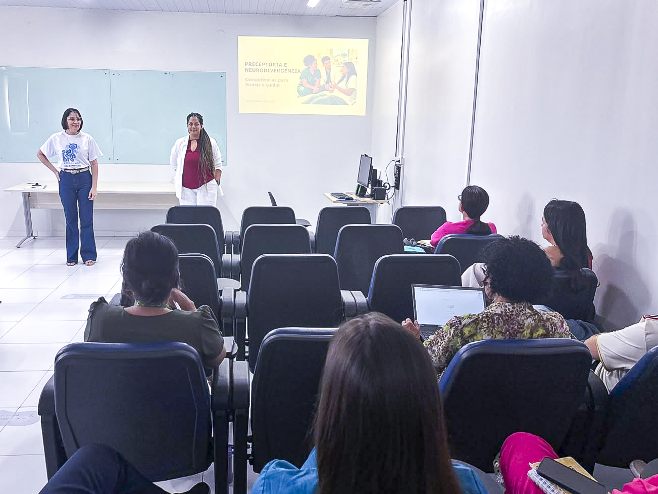 A imagem registra uma apresentação ou palestra em sala de aula / auditório, onde duas palestrantes estão de pé à frente do quadro branco e da tela de projeção. A tela exibe o título "Preceptoria e Neurodivergência" acompanhado por uma ilustração. Os participantes e ouvintes estão dispostos nas poltronas voltados para as palestrantes.