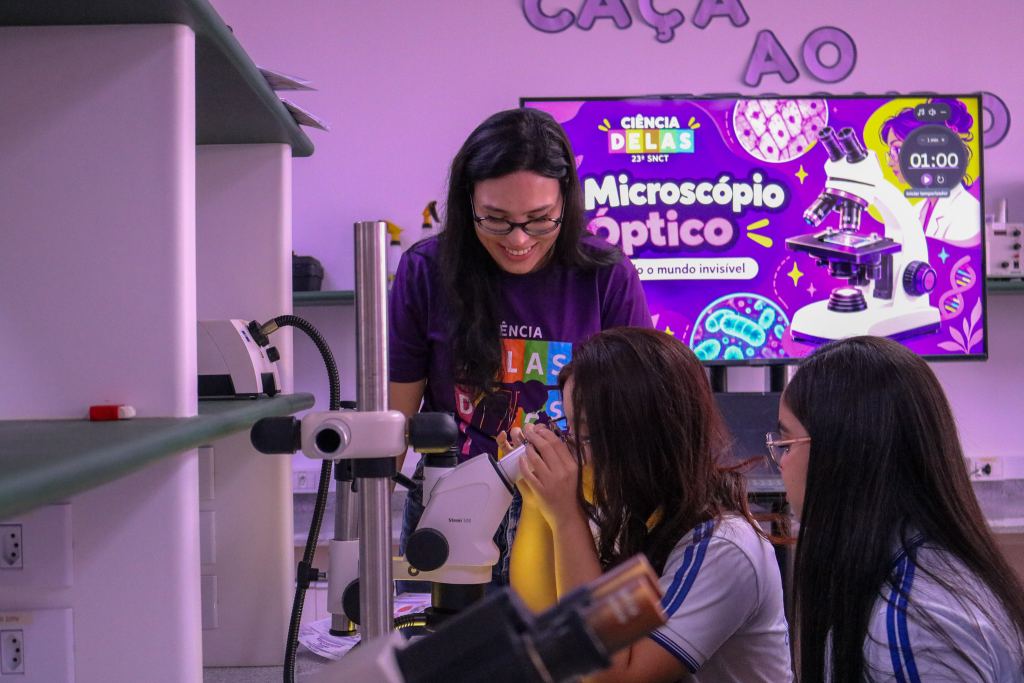 Fotografia em ambiente de laboratório com iluminação em tons de roxo e rosa. Ao fundo, vê-se uma tela exibindo a arte do projeto "Ciência Delas - 23ª SNCT", com o título "Microscópio Óptico". No centro, uma mulher adulta de cabelos escuros e óculos, vestindo uma camisa roxa, sorri enquanto orienta uma estudante que olha através das lentes de um microscópio estereoscópico branco (Stemi 508). À direita, outra jovem de cabelos longos e óculos observa a atividade atentamente. Em primeiro plano, à esquerda, há prateleiras e partes de equipamentos de laboratório desfocadas.