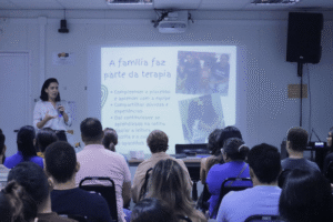 A imagem registra uma palestra ministrada no auditório do ISD sobre a importância do apoio familiar na reabilitação auditiva. A palestrante aparece de pé à esquerda, conduzindo a apresentação para o público atendo nas fileiras de cadeiras. Ao fundo, o slide projetado traz o título "A família faz parte da terapia".