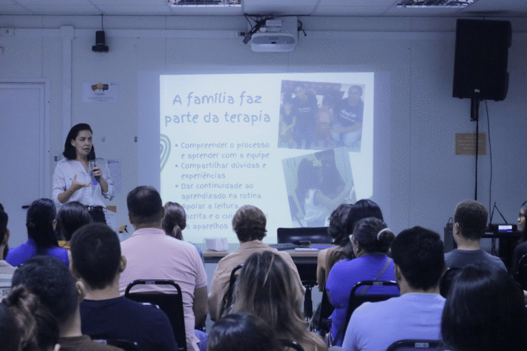 A imagem registra uma palestra ministrada no auditório do ISD sobre a importância do apoio familiar na reabilitação auditiva. A palestrante aparece de pé à esquerda, conduzindo a apresentação para o público atendo nas fileiras de cadeiras. Ao fundo, o slide projetado traz o título "A família faz parte da terapia".