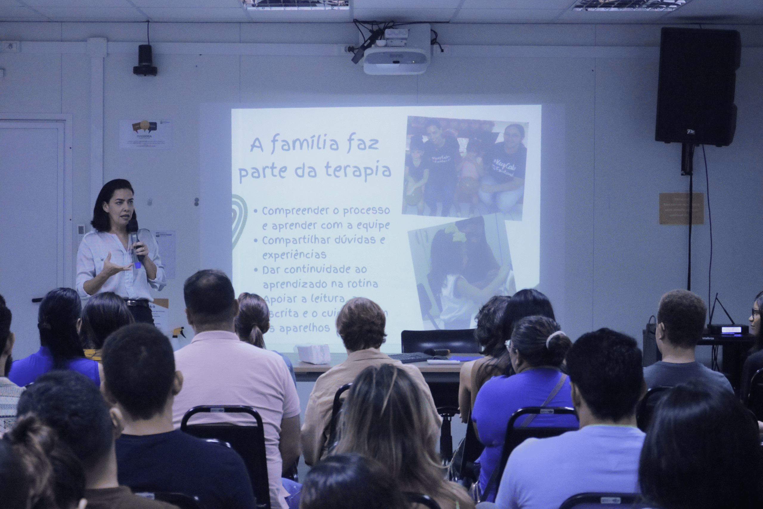 A imagem registra uma palestra ministrada no auditório do ISD sobre a importância do apoio familiar na reabilitação auditiva. A palestrante aparece de pé à esquerda, conduzindo a apresentação para o público atendo nas fileiras de cadeiras. Ao fundo, o slide projetado traz o título "A família faz parte da terapia".