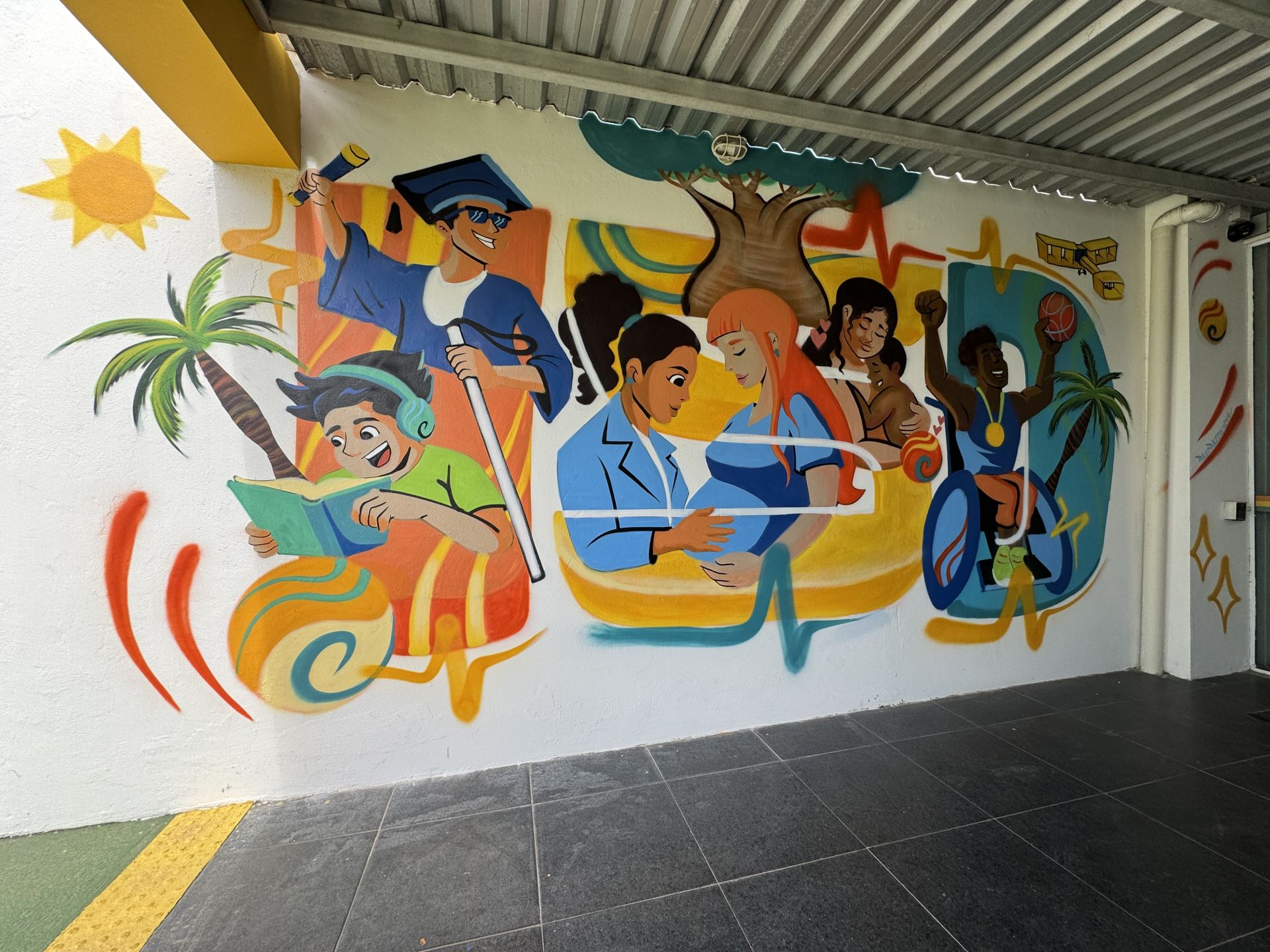 Fotografia frontal de um mural de arte urbana recém-concluído em uma parede branca sob uma cobertura metálica. A pintura retrata cenas de inclusão, diversidade e saúde, incluindo um jovem negro de óculos escuros e capelo de formatura segurando um diploma e uma bengala longa, um jovem lendo um livro, profissionais de saúde cuidando de uma gestante, e um atleta negro em uma cadeira de rodas levantando o braço com uma medalha.