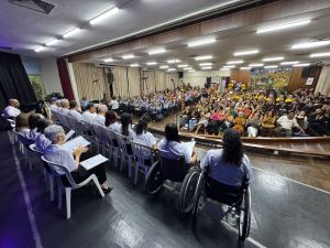 A fotografia registra a apresentação do Coral ISD a partir de um ângulo elevado no palco de um auditório lotado. Em primeiro plano, os integrantes do coral estão dispostos de costas para a câmera em duas fileiras voltadas para o público: a fileira da frente conta com coralistas sentados em cadeiras plásticas brancas e, na ponta direita, duas mulheres em cadeiras de rodas de armação escura. Os participantes vestem camisetas brancas com o logotipo da instituição e seguram pastas ou folhas de papel com as partituras nas mãos. Ao fundo, a plateia ocupa as poltronas do auditório sob iluminação de lâmpadas tubulares no teto de madeira, acompanhando a apresentação com palmas. No canto superior direito do auditório, observa-se um painel artístico colorido fixado na parede de madeira.