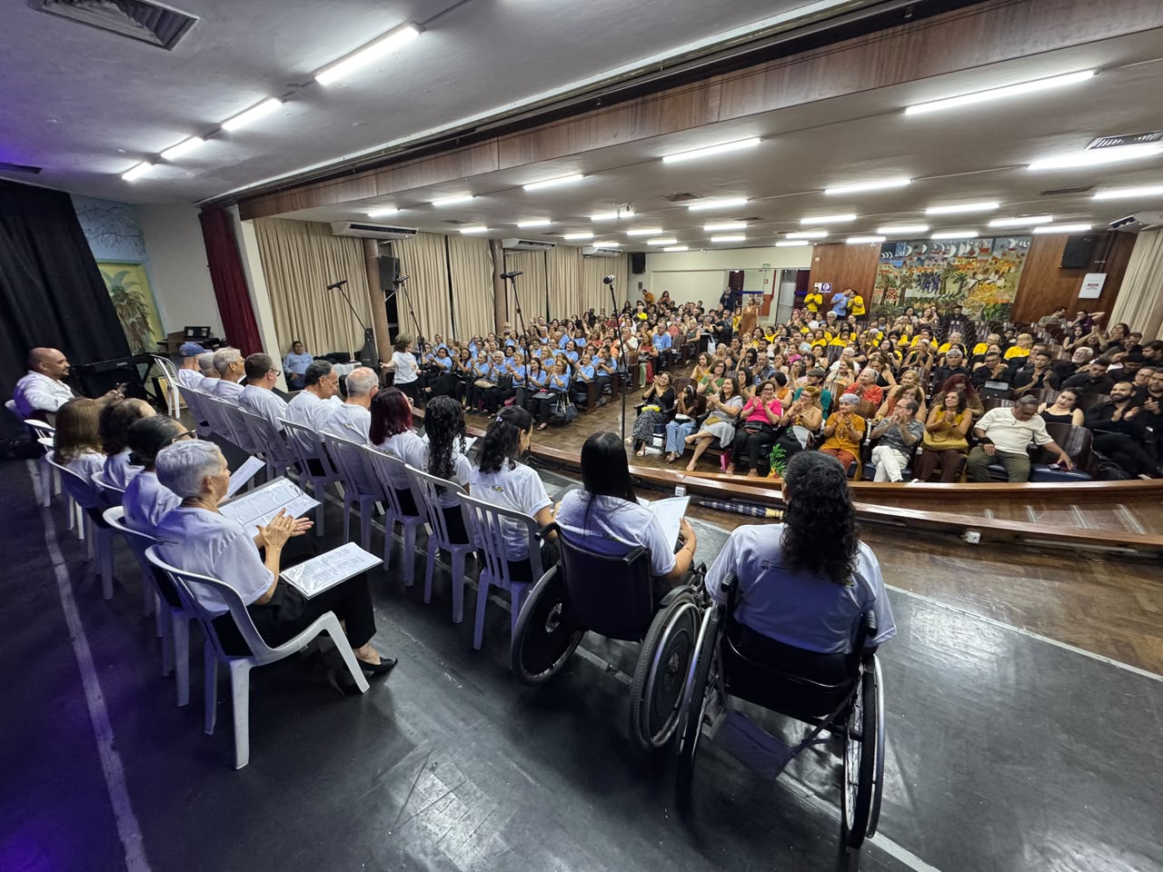 A fotografia registra a apresentação do Coral ISD a partir de um ângulo elevado no palco de um auditório lotado. Em primeiro plano, os integrantes do coral estão dispostos de costas para a câmera em duas fileiras voltadas para o público: a fileira da frente conta com coralistas sentados em cadeiras plásticas brancas e, na ponta direita, duas mulheres em cadeiras de rodas de armação escura. Os participantes vestem camisetas brancas com o logotipo da instituição e seguram pastas ou folhas de papel com as partituras nas mãos. Ao fundo, a plateia ocupa as poltronas do auditório sob iluminação de lâmpadas tubulares no teto de madeira, acompanhando a apresentação com palmas. No canto superior direito do auditório, observa-se um painel artístico colorido fixado na parede de madeira.