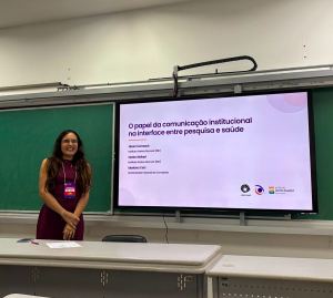 Mulher de cabelos longos e escuros, sorrindo, vestindo um vestido sem mangas na cor vinho e usando um crachá, está em pé à esquerda em uma sala de aula. À direita dela, há uma lousa verde e uma grande tela digital exibindo uma apresentação com o título "O papel da comunicação institucional na interface entre pesquisa e saúde", além de nomes de pesquisadores e logotipos das instituições envolvidas (Unicamp e Instituto Santos Dumont).